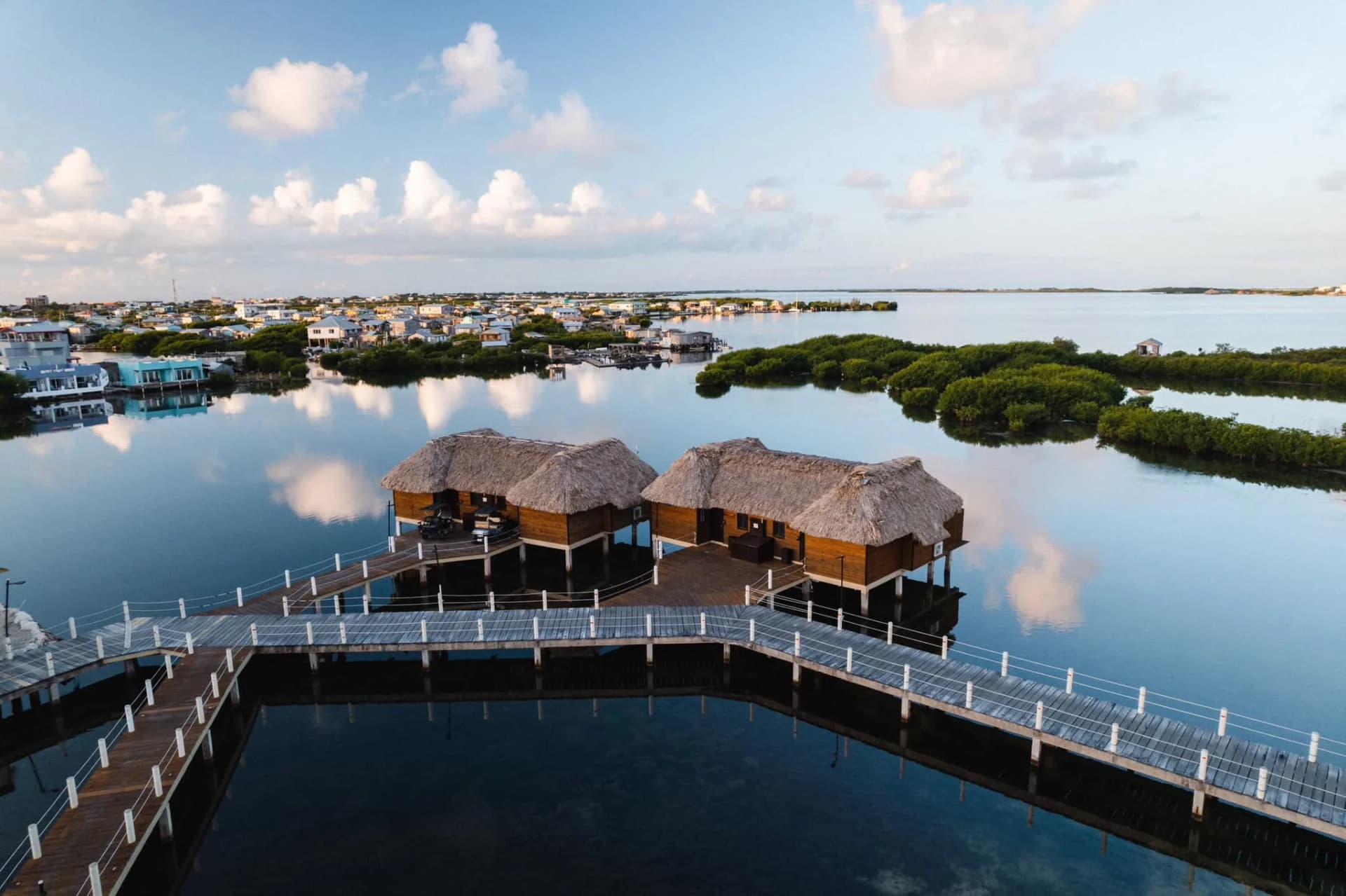 Two thatched overwater cabanas on a white-railed boardwalk, reflected in a mirror-calm lagoon.
