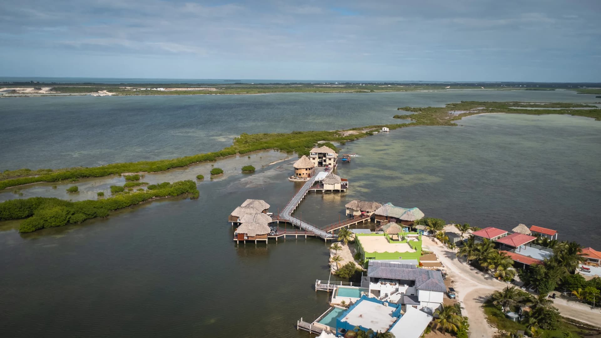 Aerial view of Lina Point Overwater on Ambergris Caye, Belize: a long boardwalk over the lagoon to thatched overwater buildings, with mangroves and the Caribbean beyond.