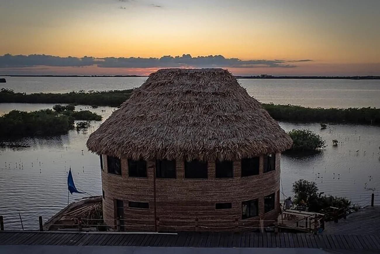The Mayan's thatched roundhouse silhouetted against a gold sunset over the lagoon.