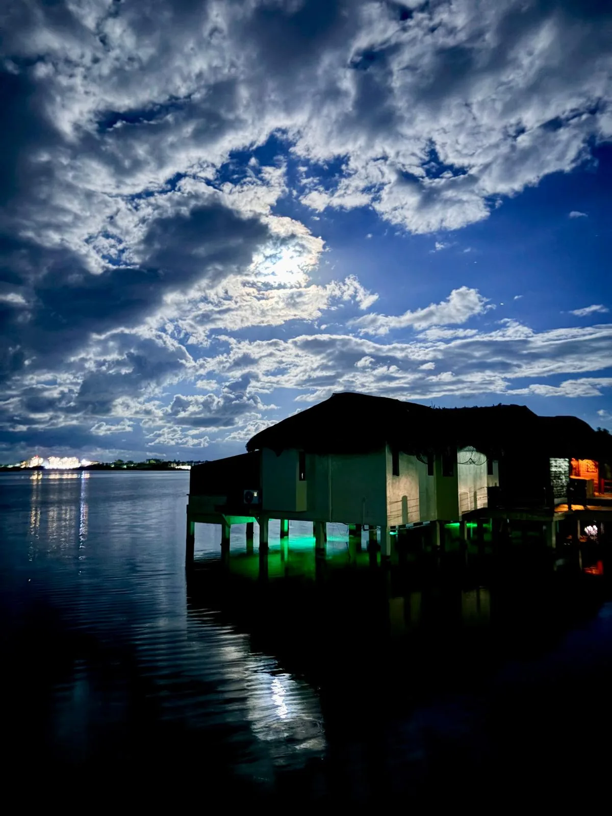 An overwater cabana at night under moonlit clouds, green lights glowing beneath it on the water.